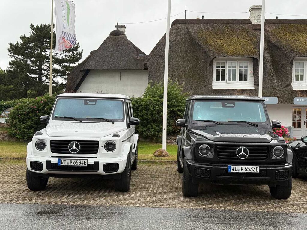 mercedes-g-580-fahrbericht-sylt Der neue Mercedes G 580 EQ in weiss und Schwarz vor einem Redgedeckten Gebäude stehen auf einem Parkplatz